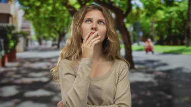 Kadın endişeli bir bakışla eline bastırır ve bir akıllı telefonu kavrar; sarışın genç ziyaretçi aramadan önce endişeli bir şekilde parkın açık alanında durur.