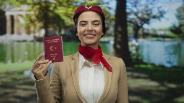 Üniformalı İspanyol bir uçuş görevlisi, elinde Türk pasaportu, güneşli bir parkta duruyor, arka planda dingin bir göl ve ağaçlar var..