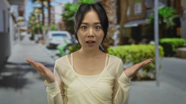 Genç Çinli kadın güneşli bir caddede dikiliyor. Arka planda palmiye ağaçları ve şehir sokaklarında park edilmiş arabalarla sürpriz yapıyor..