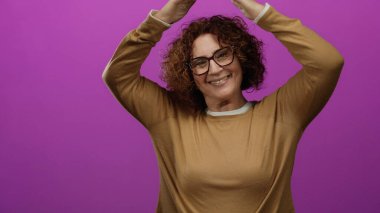 Çatı jesti yapan kadın, elleri parlak pembe arka planda, gözlük ve kahverengi süveter giyerken neşeli bir ifade sergiliyor, evi ve mutluluğu gösteriyor..