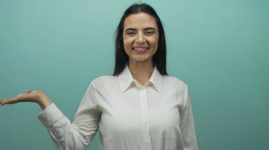 Genç İspanyol kadın gülümsüyor ve elini mavi bir duvara doğru uzatıyor, özgüven ve duruş ile sunum ya da tanıtım öneriyor.