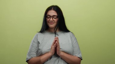 Sarı arka plan üzerinde sakince meditasyon yapan, gözlüklü, gri gömlekli, elleri birarada sakin ve sakin tavırları olan bir kadın..