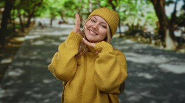 Hardal süveter ve şapkayla gülümseyen bir kadın, yemyeşil ağaçlarla dolu güneşli bir parkta mutlu poz veriyor..