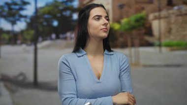 Genç İspanyol kadın güneşli bir sokakta dikiliyor. Mavi giyinmiş, ağaçların ve binaların arka planında huzurlu şehir hayatını yansıtıyor..