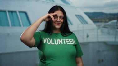 Gezgin gemisinde gönüllü olan bir kadın jest yaparken gülümsüyor, açık havada yeşil gömlek giyiyor..