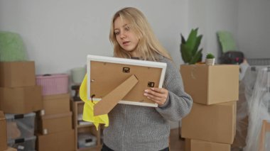 Oturma odasındaki resim çerçevesini temizleyen genç bir kadın, taşınan kutularla çevrili, yeni bir ev ortamının rahat bir sahnesini tasvir ediyor..