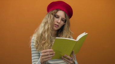 Stüdyoda kitap okuyan bir kadın, kırmızı bereli ve gözlüklü genç ve sinirli bir sarışını sergiliyor..