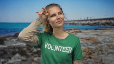 Kumsaldaki sarışın gönüllüye terini silen, güneşli okyanus göğünün altında yeşil tişört giyen kayalık önplanlı ve yorgun görünen bir kadının portresi..