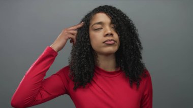 Kadın gri stüdyoda başını yukarı kaldırır. Kırmızı üst ve kıvırcık saçlar anlamlı yüz ve düşünceli bakışlar gösterir..
