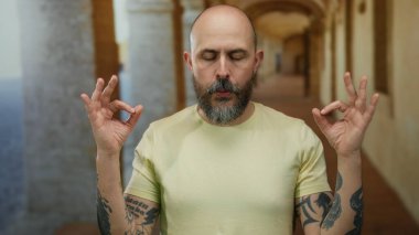 Eski bir üniversitede meditasyon yapan bir adam gözleri kapalı ve dövmeleri görünür, sakin ve huzurlu bir atmosfer yaratıyor..
