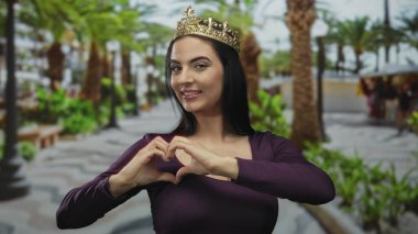 Taçlı bir kadın, güneşli bir parkta, etrafı palmiye ağaçlarıyla çevrili bir şekilde, dışarıda eğlenmeyi ve kutlamayı simgeleyen bir kalp hareketi yapıyor..