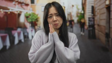 Açık hava restoranının terasında elleri dua pozisyonunda olan genç Çinli bir kadın, bir sokak ortamında duygu ya da özür diliyor, bulanık yayalar ve binalarla çevrili..