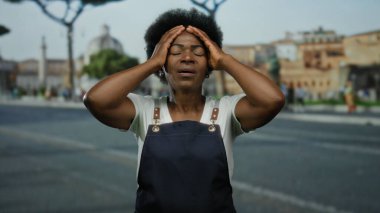 Önlüklü bir kadın şehir caddesinde hayal kırıklığı içinde başını tutuyor. Bulanık binalar ve ağaçlarla şehir zeminine karşı stresi dile getiriyor..