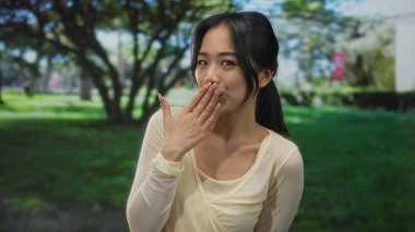 Parktaki genç kadın mutlulukla gülümsüyor ve jestler yapıyor, yemyeşil ağaçlar ve çimlerle çevrili, dingin ve canlı bir atmosfer sergiliyor..