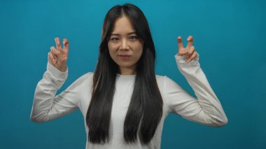 Çinli kadın mavi arka plan üzerinde tırnak işareti yapıyor, beyaz bluz giyerken konsept veya mizah ifade ediyor; genç ve kendinden emin ifade.