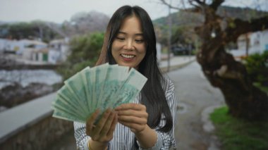 Açık havada gülümseyen genç bir kadın deniz kenarındaki bir gezinti alanında elinde Çin Yuan banknotları ile huzurlu bir ortamda ağaçların ve binaların arkasında..