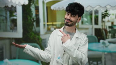 Beyaz önlüklü genç adam, arka planda yapraklar ve beyaz tentelerle restoranın terasında el kol hareketi yapıyor. Hoş bir gülümsemeyle misafirperver bir tavır sergiliyor..