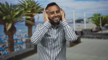 Stüdyoda kulaklık tutan bir adam plaj arkaplanı ve palmiye ağaçları ile dinlerken gülümsüyor; keyif rahatlaması.
