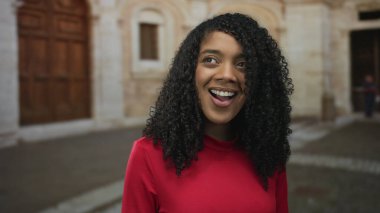 Kırmızı tişörtlü kadın eski kasabanın parke taşlı sokağında kıvırcık saçlarıyla gülümseyen yumuşak güneş ışığıyla yıkanan; mutlu keşif.