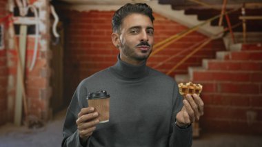Waffle ısıran ve gri balıkçı yaka kazaklı kahve fincanı tutan bir adam inşaat binasının içinde; rahatlık düşkünlüğü.