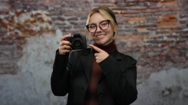 Sarışın bir kadın kamerayı işaret ediyor. Tuğla bir duvara gülümsüyor. Arkaplanda gözlük ve siyah bir ceket var..