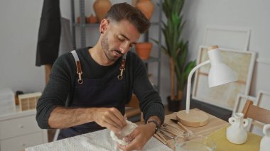 Hispanik adam parlak bir stüdyoda, ahşap bir masanın üzerinde çıplak elle kil heykeli şekillendirir; yaratıcı odaklanma.