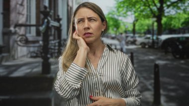 Sarı saçlı ve çizgili bluzlu bir kadın meşgul bir şehir caddesinde bina cephesine yakın kulağına bastırıyor..