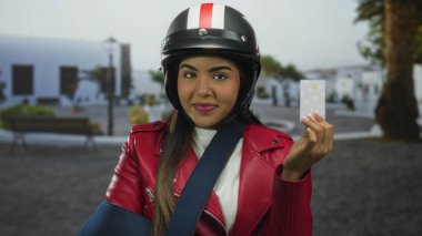 Kasklı, kırmızı ceketli, kol askısı ve kredi kartıyla sokakta elinde motosikletle gezen genç bir kadın..