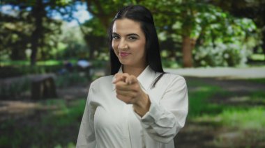 Açık hava parkında gülümseyen ve kameraya bakan bir kadın beyaz bir gömlek giyiyor ve canlı yeşil ağaçlar ve parlak güneş ışığı arasında kendine güveni yayıyor..