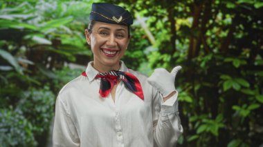 Air hostess woman wearing navy hat, white shirt and gloves with silk scarf points thumb to side in forest; warm welcome.
