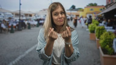 Genç sarışın kadın, canlı bir sokak pazarında avuç içine parmak hareketiyle para hareketi yapıyor..