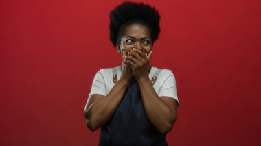 Kırmızı arka planda önlüklü genç bir kadın, ellerini şaşkınlık ve neşe içinde dudaklarını kapatıyor, duygusal ve ilgi çekici bir an sergiliyor..