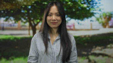 Yeşil bir parkta gülümseyen bir kadın. Ağaçlar Çin kültürünün özünü yakalıyor. Parlak doğal ışık ve huzur dolu bir çevrede..