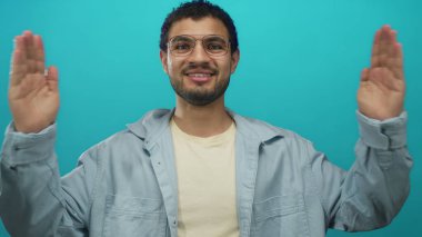 Gözlüklü sakallı adam stüdyoda turkuaz duvara karşı hafif gömlek giyerek el ele tutuşuyor; iyimserlik..