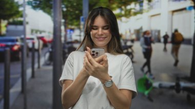 Şehir caddesinde insanlarla çevrili akıllı telefon kullanırken gülümseyen bir kadın ve güneşli bir günde açık havada günlük giysiler giymiş, odaklanmış ve rahat görünen bir kadın..