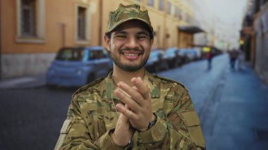 Kamuflaj üniformalı genç İspanyol adam, parlak güneş ışığı altında yoğun şehir caddesinde enerjik bir şekilde el çırpıyor..