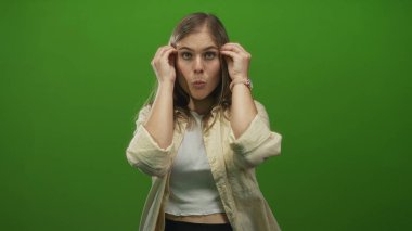 Genç sarışın kadın, parlak yeşil stüdyo setinde kameraya bakarken elleri ile yüzünü çerçeveler; şaşkınlık.
