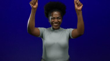 Kollarını kaldırarak kutlayan bir kadın, izole edilmiş mavi bir arka planda mutluluğu ve güveni ifade ediyor, genç bir Afro-Amerikan kadınını neşe içinde gösteriyor..