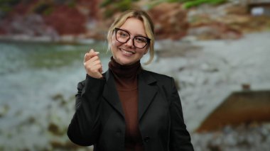 Genç bir kadın deniz kenarında gülümserken el hareketi yapıyor bulanık plaj ve okyanus geçmişiyle..