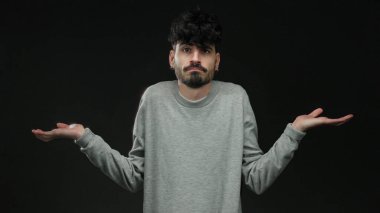 Siyah bir arkaplana karşı kafa karışıklığı içinde omuz silken, gri bir kazak giyen, kararsızlık ve belirsizlik gösteren genç bir adam yakın plan bir portrede.