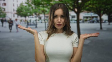 Beyaz elbiseli kadın, bulanık yayalarla meşgul şehir caddesinde elleri omuz silkerek çıplak geziyor; belirsizlik.