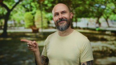 Sakallı ve dövmeli kel bir adam güneşli parkta etrafı yemyeşil ağaçlarla çevrili, arkadaşlığı ve gelişigüzel tarzını tasvir ediyor..