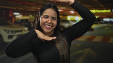 Latin asıllı genç fotoğrafçı tarafından park yerinde yakalanan kadın yüz çerçevesi. Kapalı alanda gülümsüyor. Elleri hafifçe öne çıkıyor..