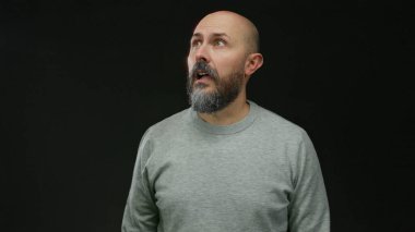 Sakallı bir adam, izole edilmiş siyah bir arkaplana şaşkınlıkla bakıyor gri bir süveter giyiyor. Hislerini minimalist ortamda ifade ediyor..
