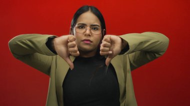 Kırmızı arka planda, gözlüklü ve ceketli bir kadının onaylamadığını belirten bir kadın, izole stüdyo ortamında olumsuz duygular ve ciddi ifadeler sergiliyor..