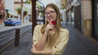 Gözlüklü bir kadın gülümsüyor ve iki eliyle kırmızı kalp kartı tutuyor. Gün ortasında güneşli şehir caddesinde..