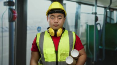 Güvenlik yeleği ve kasketi takmış, şehir gelişimini ve toplu taşıma planlarını sembolize eden Asyalı genç bir adam otobüsün içinde duruyor..