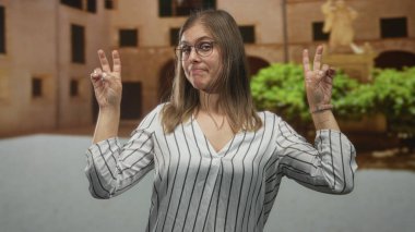 Sarışın genç bir kadın tarihi bir kasaba binasında sırıtıyor ve parmaklarıyla tırnak işareti yapıyor..
