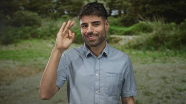 Açık mavi tişörtlü genç İspanyol adam yeşil bir parkta sağ eliyle 