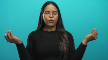 Kadın sakin bir şekilde meditasyon yapıyor gözleri kapalı ve elleri mudra pozisyonunda izole edilmiş yeşil bir arka plan üzerinde, sakin bir ortamda gözlük ve siyah kıyafetler giyiyor..